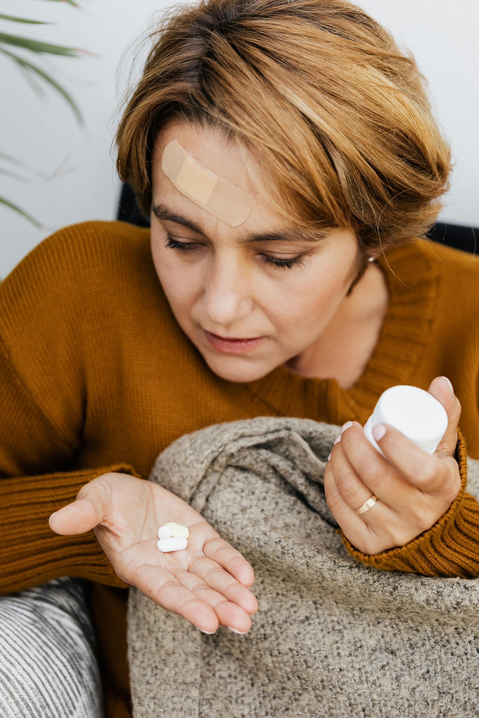 Woman looking at pills in her hand, bandage on her forehead, holding a pill bottle.