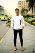 Man standing on a sidewalk, hands in pockets, smiling. He wears a white sweater, black pants, and tan shoes. City street backdrop.