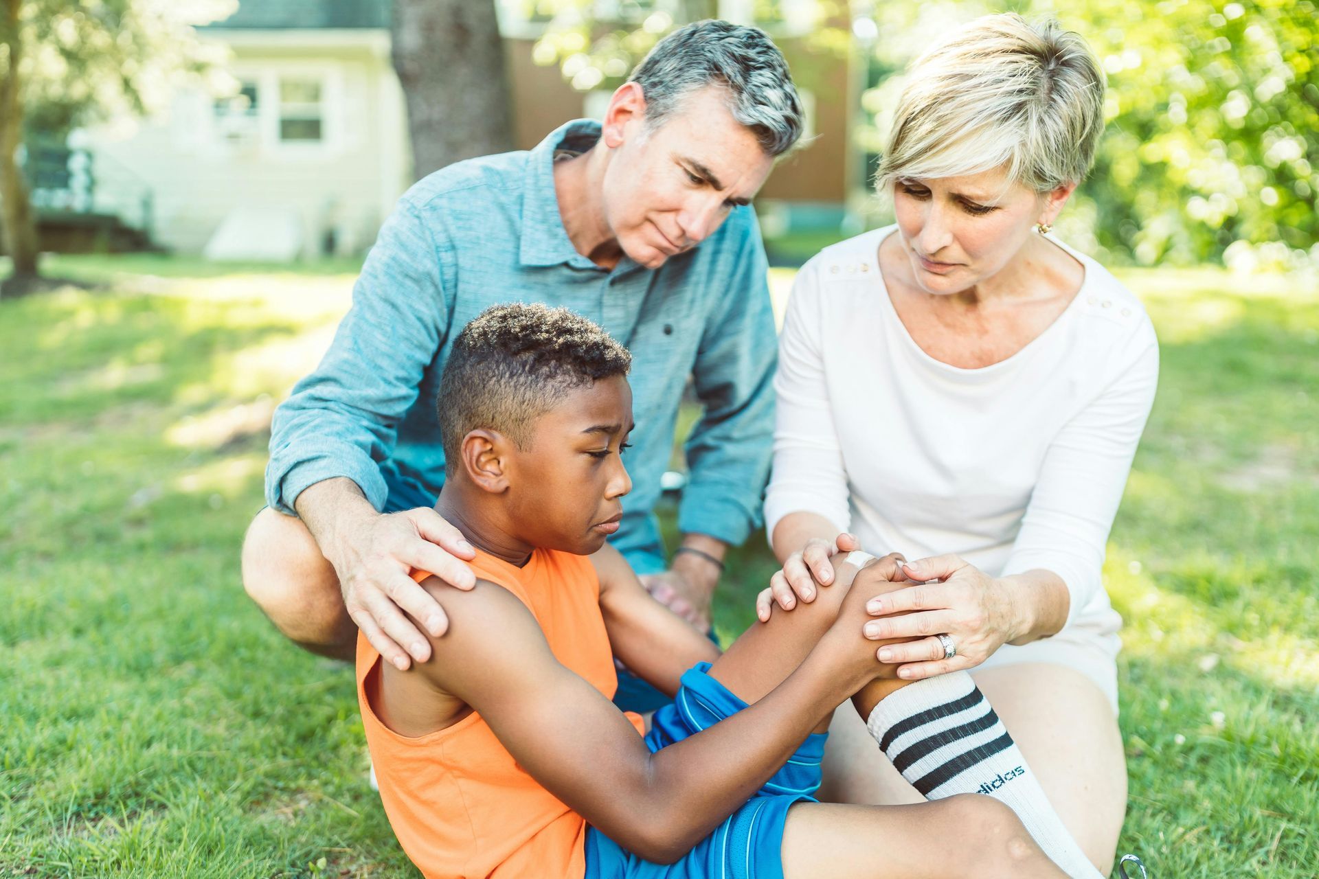 Family comforts a child with an injured knee while sitting on grass outside.