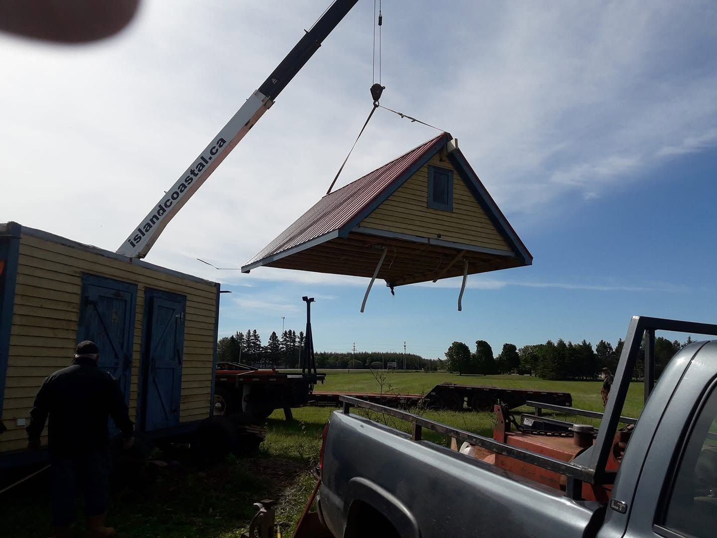 Crane lifting a small yellow house roof over a truck in a rural field