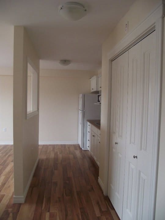 Empty hallway with wood floors, white walls, closet doors, and a kitchen area at the far end.