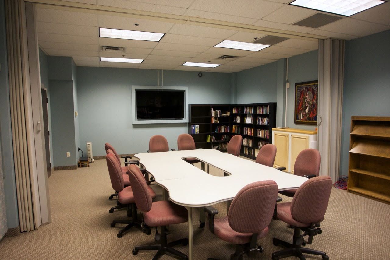 Conference room with oval table, pink chairs, wall-mounted TV, bookshelf, and curtains.