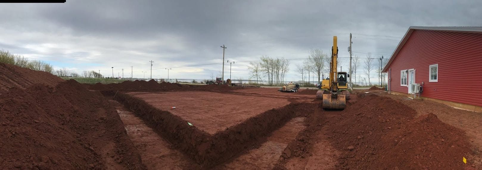 Rural construction site with red dirt trench, excavator, and a red building under a cloudy sky
