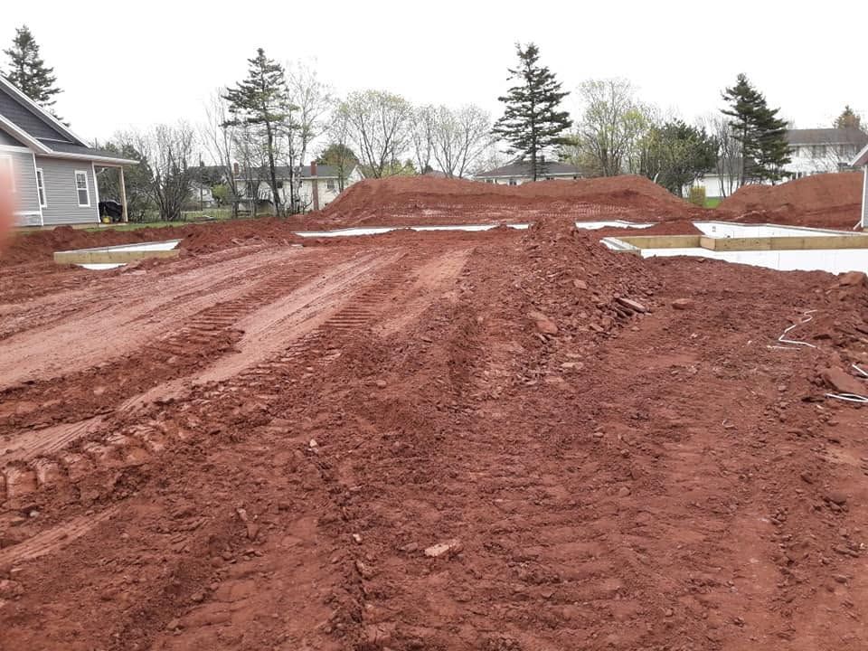 Rutted red dirt construction site with mounds of earth beside houses and trees