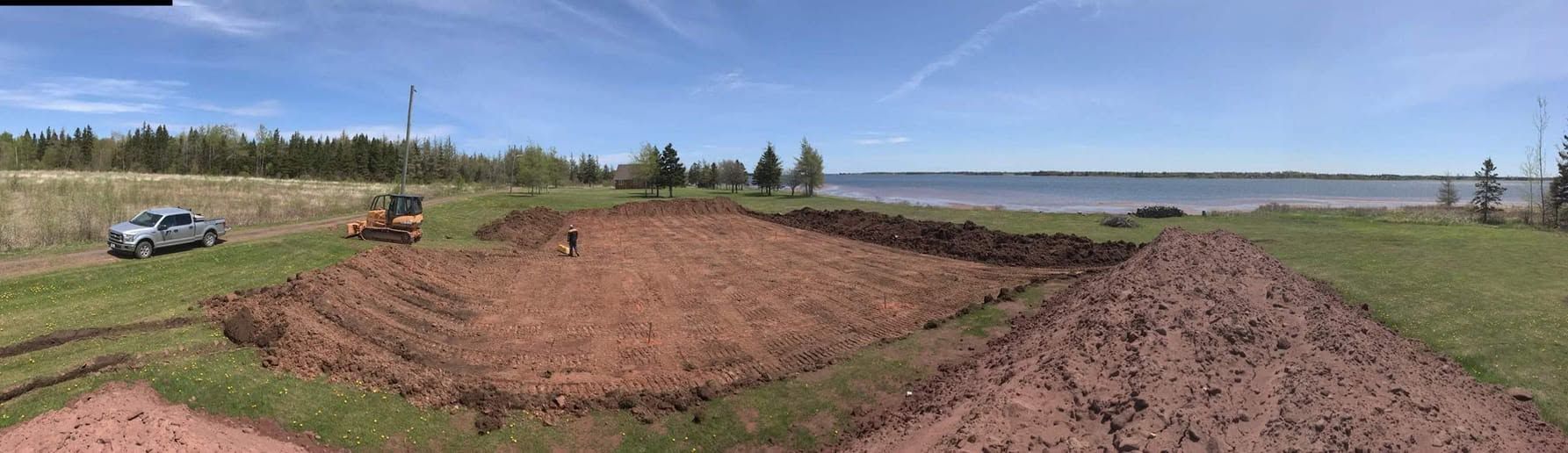 Wide dirt mound under construction in a grassy field, with a small vehicle and trees under a blue sky.