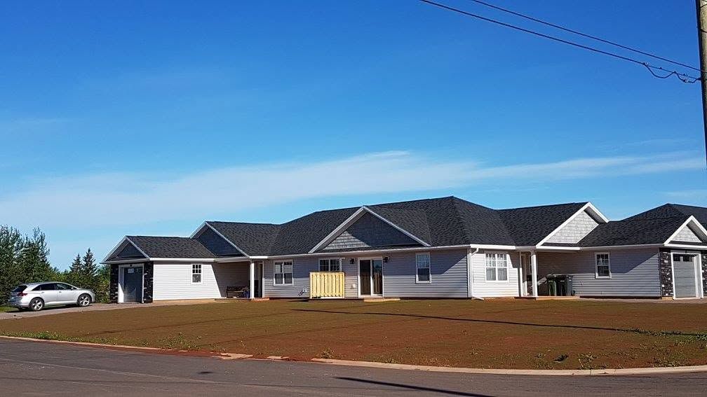 Single-story suburban houses with gray roofs under a clear blue sky, beside a dirt road.