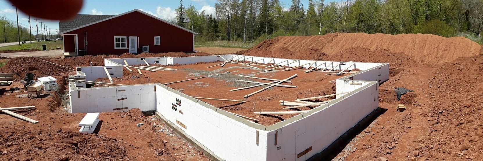 Partially built white foundation on a dirt lot with a red barn-like house in the background