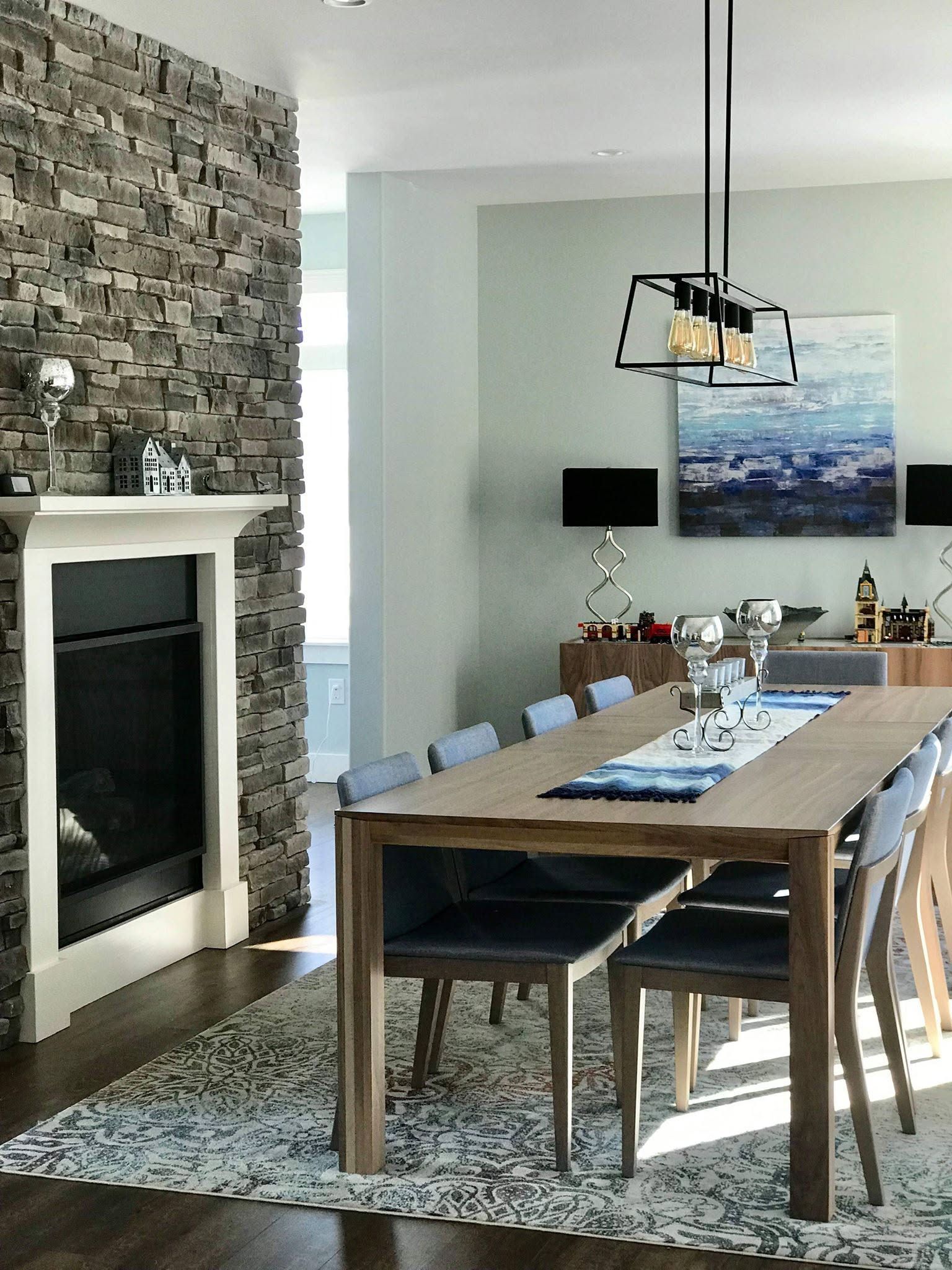 Modern dining room with stone fireplace, wood table, patterned rug, and landscape wall art.