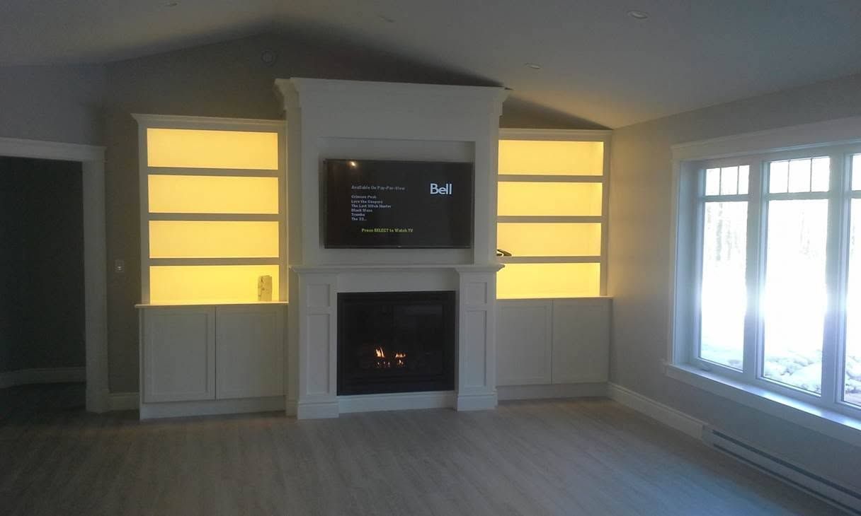 Living room with built-in fireplace, wall-mounted TV, and lit yellow cabinets beside large windows.