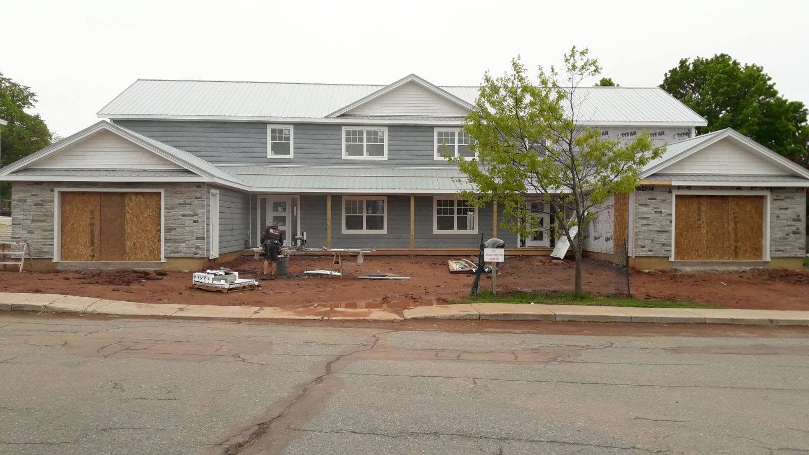 Large gray house with a metal roof, front porch, and two garage doors, set behind a bare yard and street.