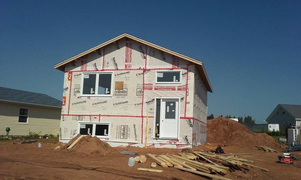 Partially built house wrapped in insulation, sitting on a dirt lot under a clear blue sky
