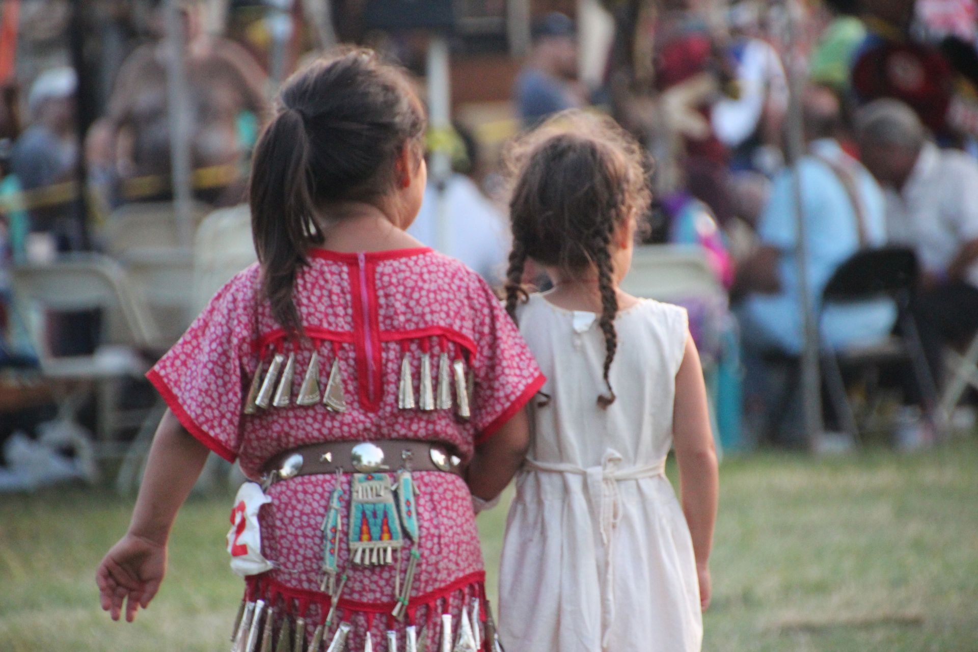 Two young girls, one in a red beaded dress, the other in white, hold hands, at a gathering.