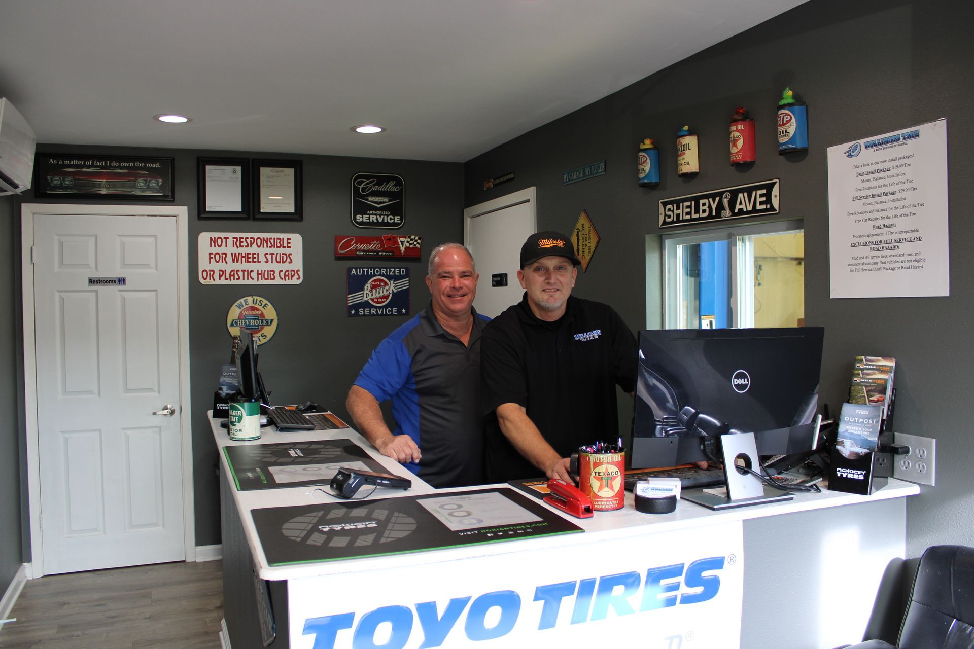 Two men standing behind a Toyo Tires counter in a shop office, with a desk, computer, and wall signs.