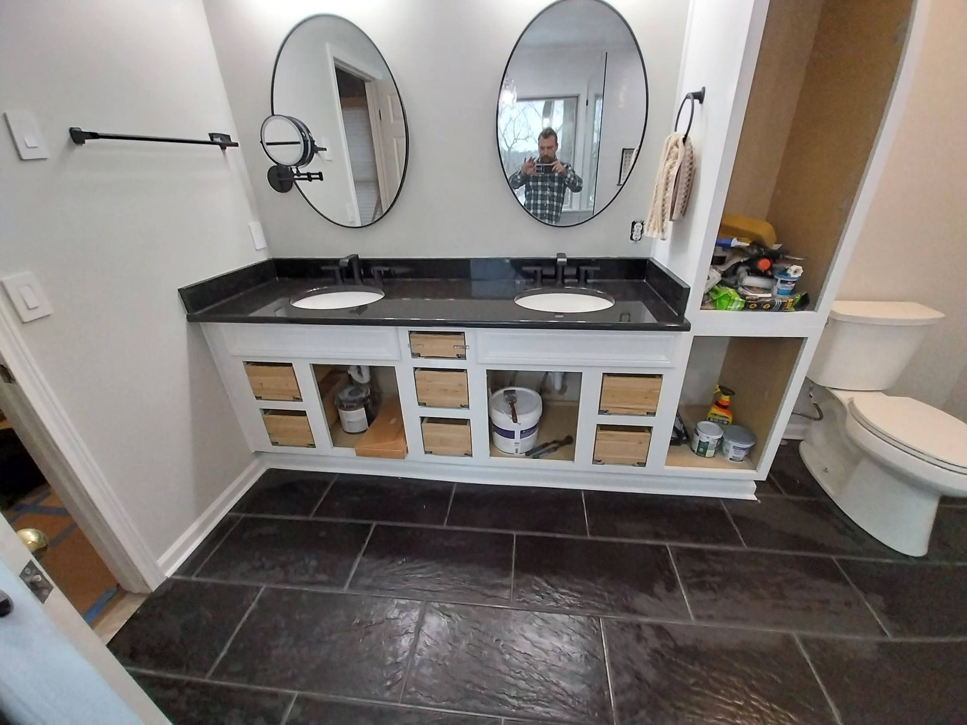 Bathroom vanity with two oval mirrors, black countertop, and dark tile floor.