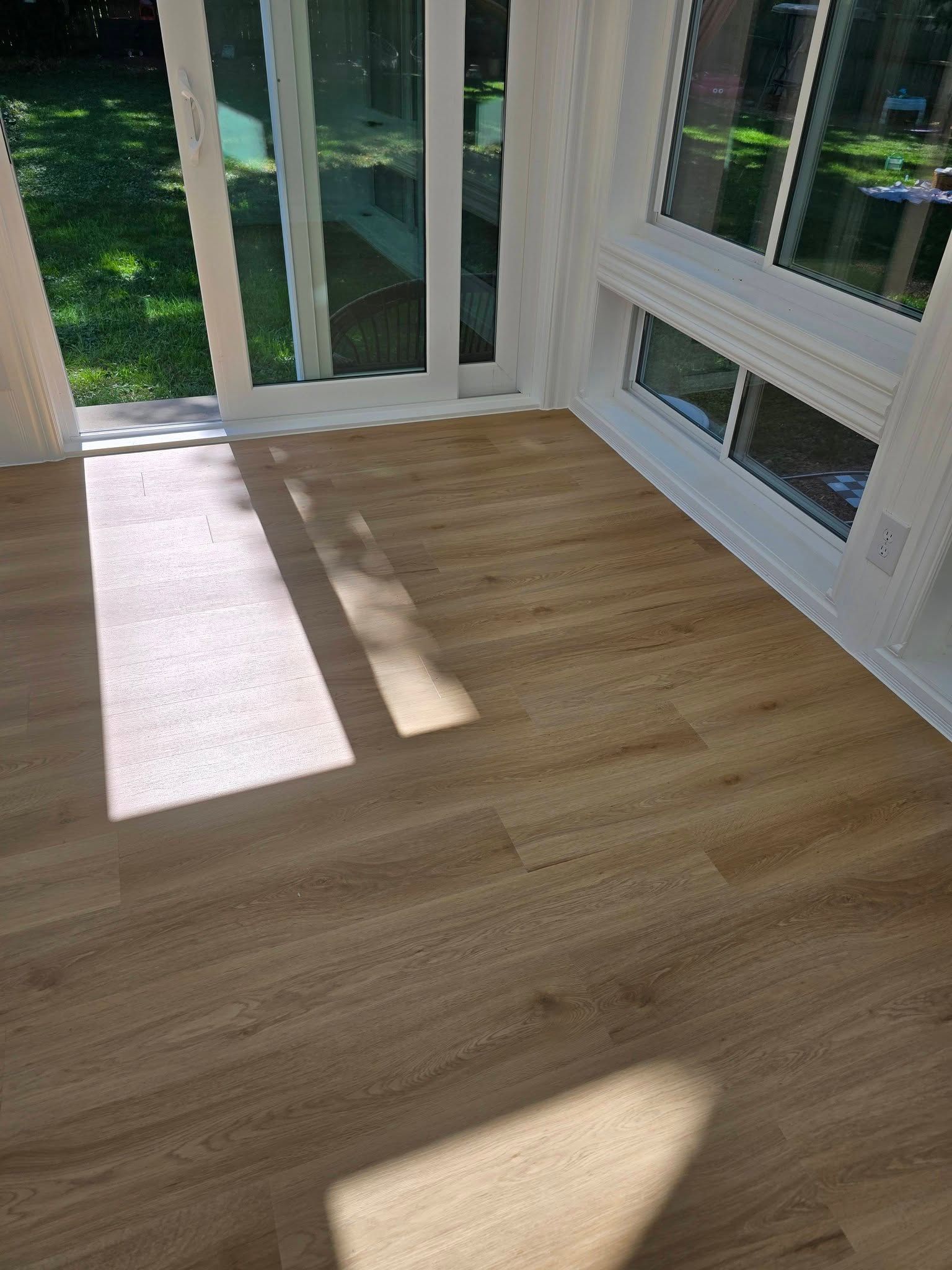 Sunlit empty room with light wood floor, white walls, and large glass doors and windows.