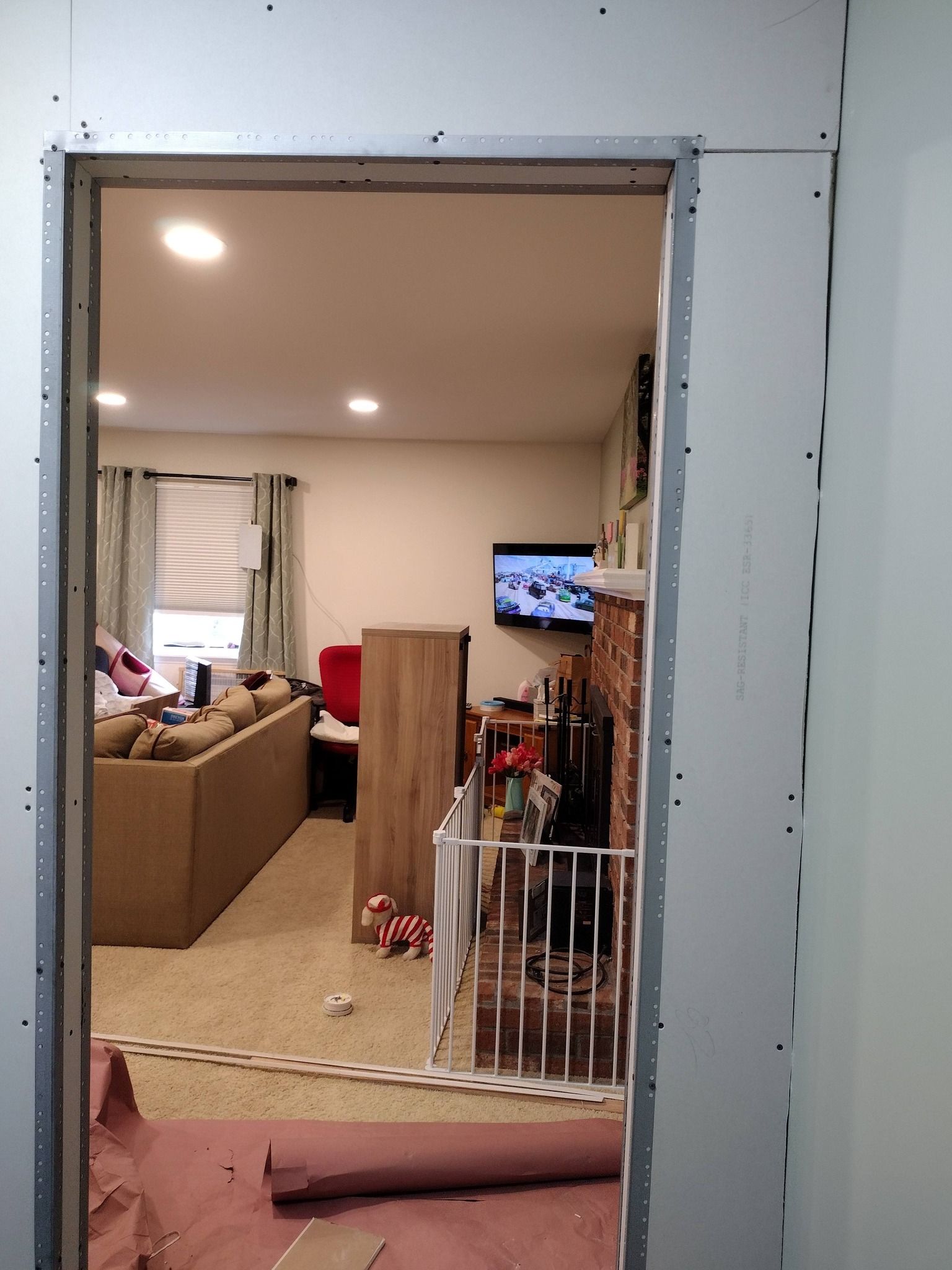 Living room seen through a doorway, with beige furniture, a TV on the wall, and a baby gate in front of a hallway.