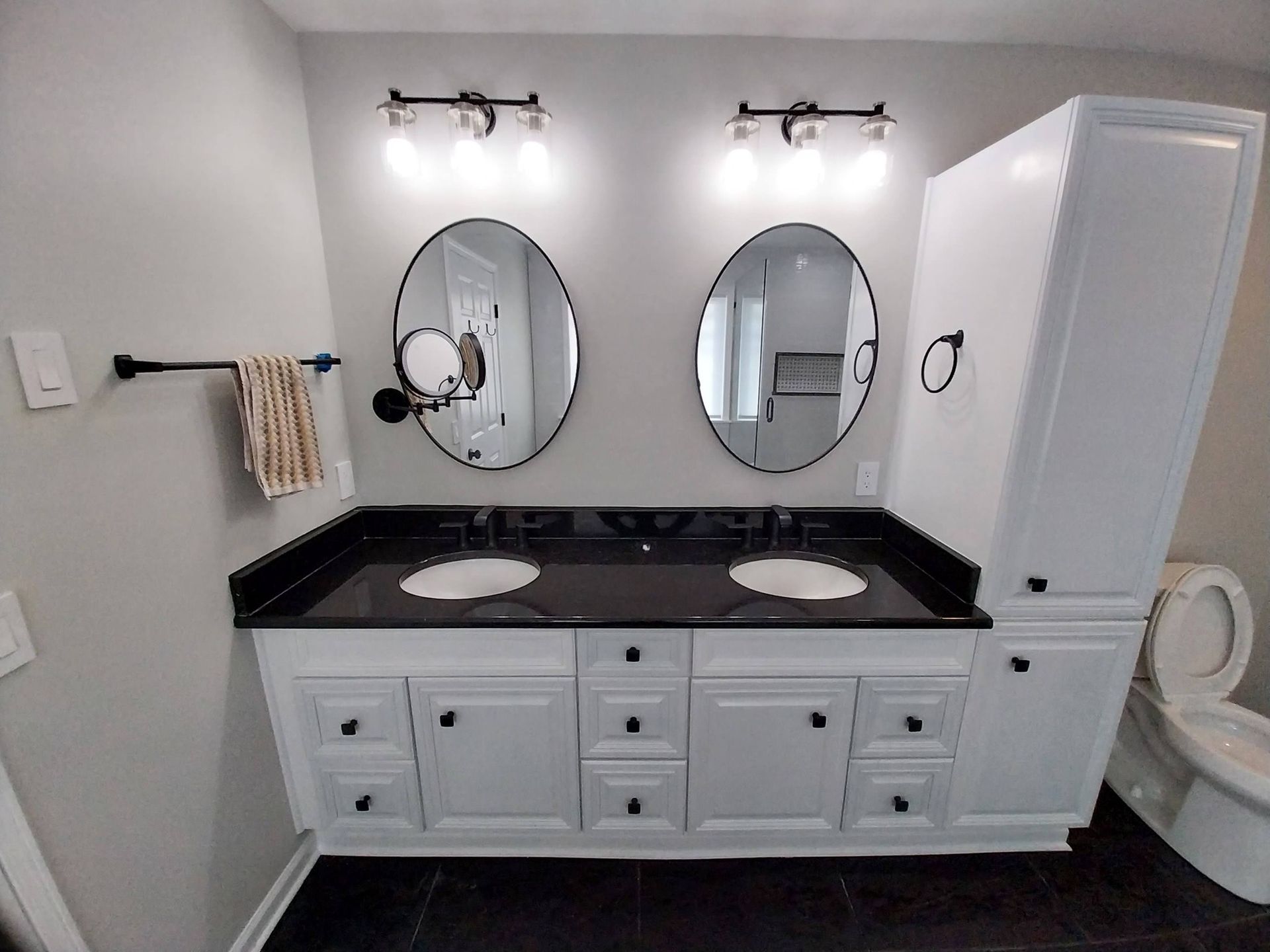 Double vanity bathroom with two oval mirrors, black countertop, white cabinets, and wall lights