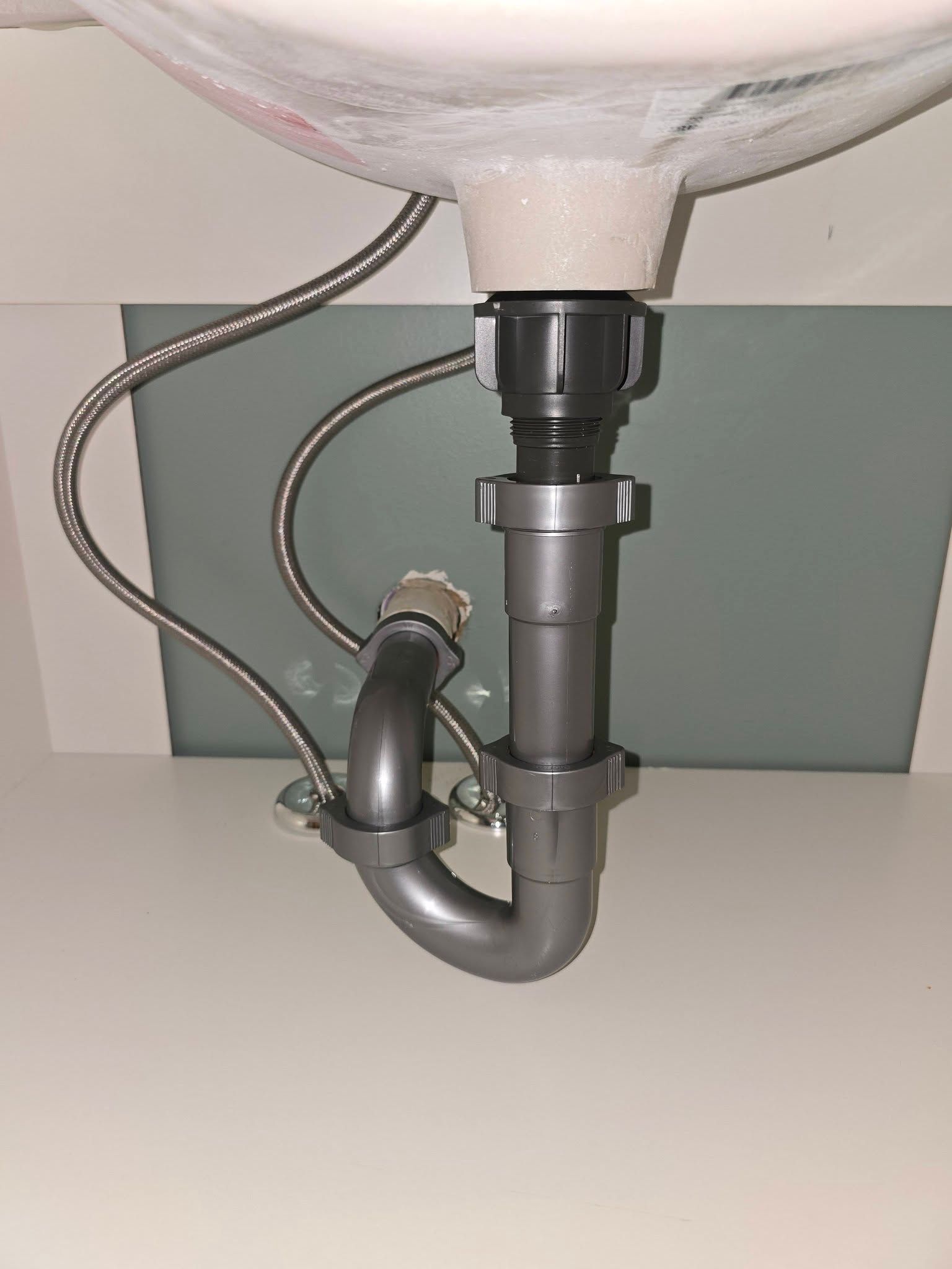 Under-sink bathroom plumbing with chrome drain pipes and a flexible hose against a gray wall