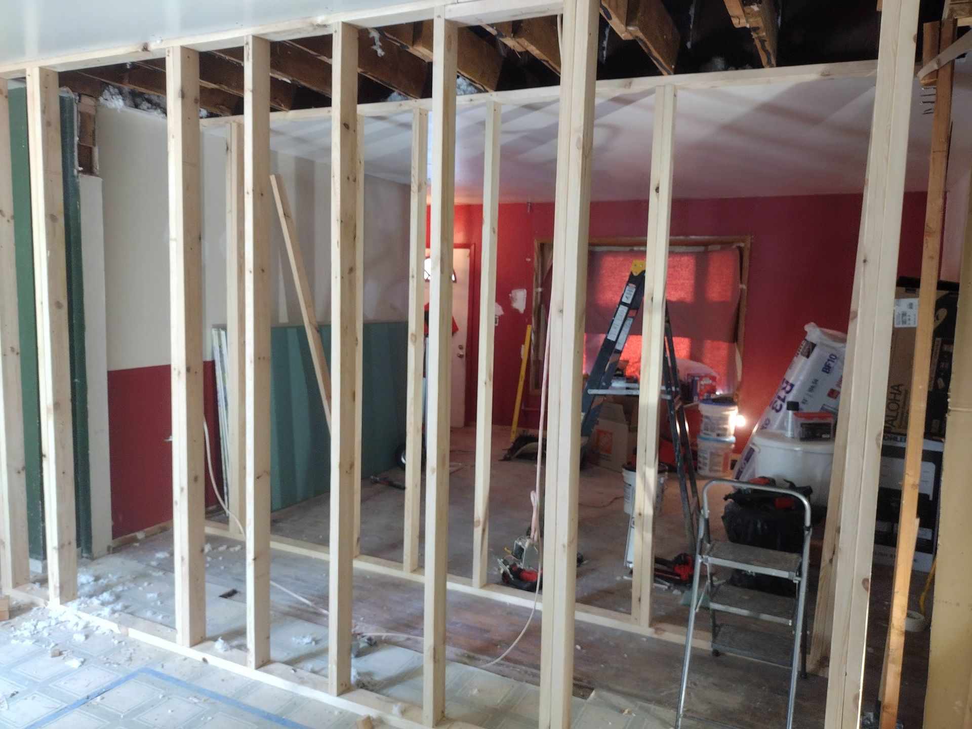 Framed interior under renovation with exposed studs, red walls, ladders, and construction debris on the floor