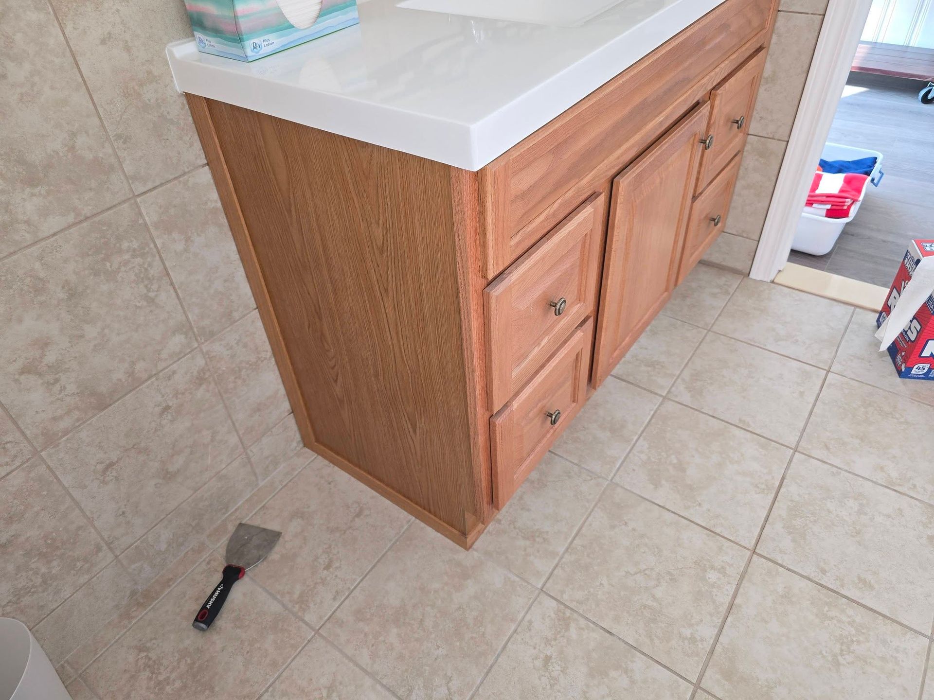 Wooden bathroom vanity with white countertop on beige tile floor, with a putty knife nearby