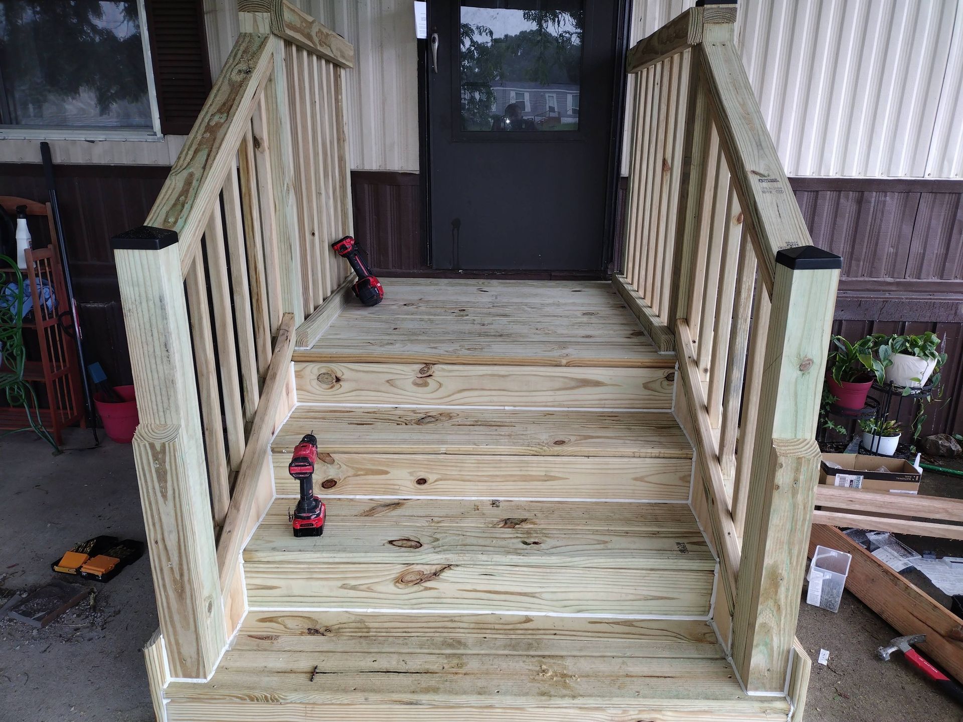 Wooden porch ramp with railings leading to a dark front door, with two red cordless drills on the steps.