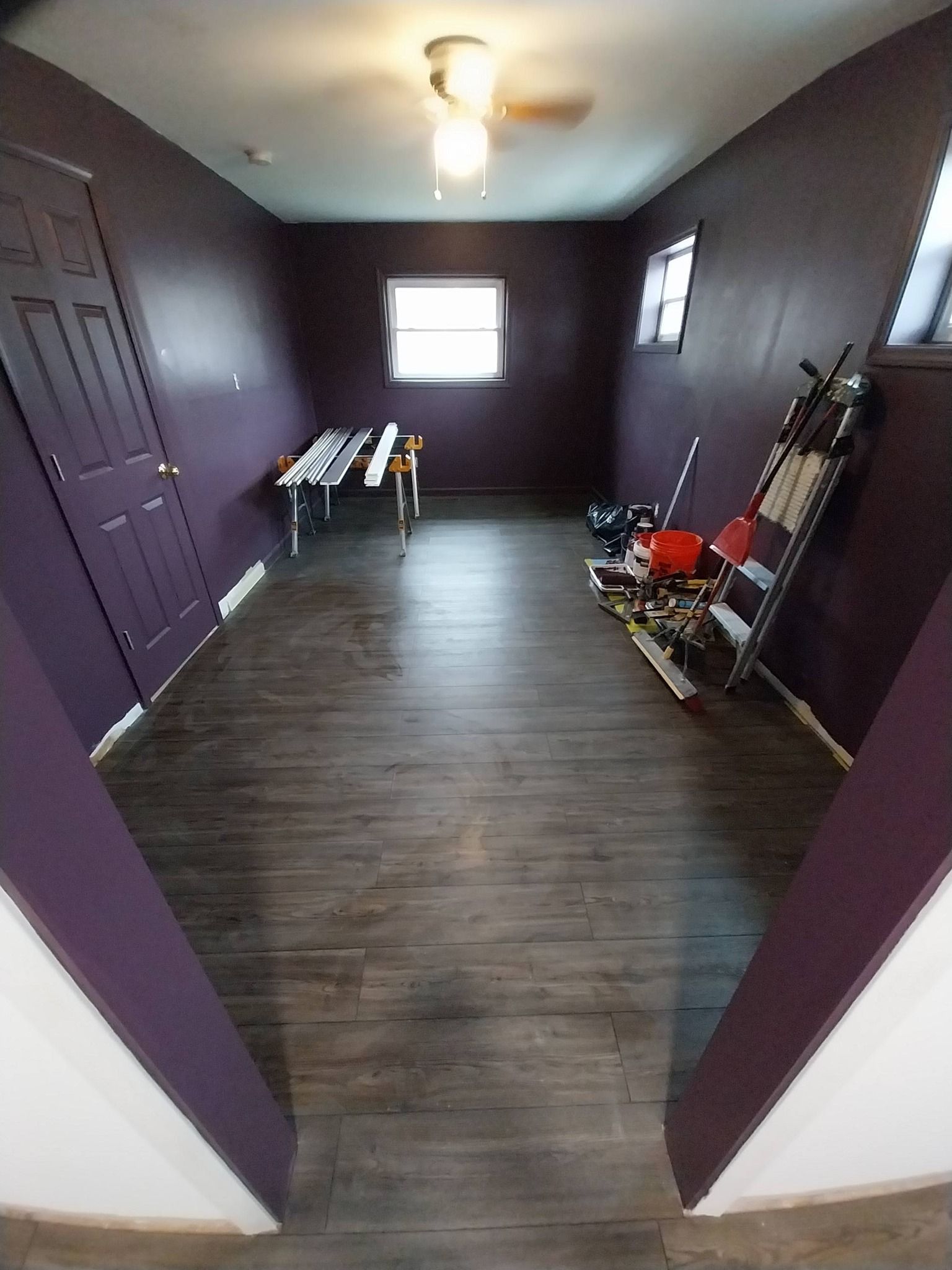 Empty purple-walled room with dark wood floor, window, ceiling light, and folding tables and chairs.