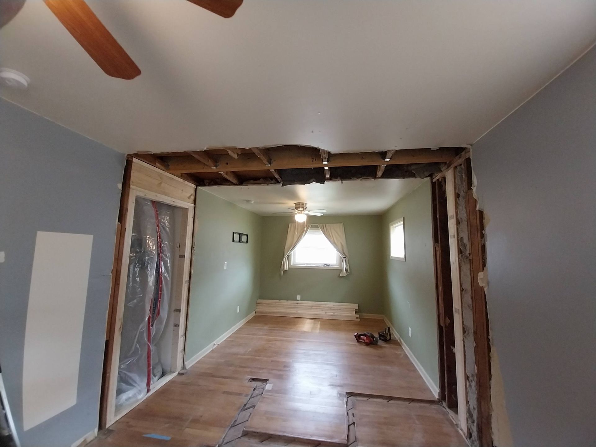 Partially gutted room with exposed framing, light green walls, hardwood floor, and a window at the far end