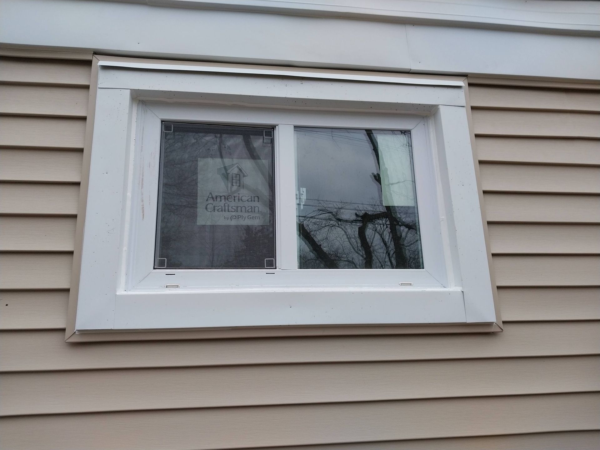 White-trimmed window on beige siding with a partially open pane and reflected tree branches