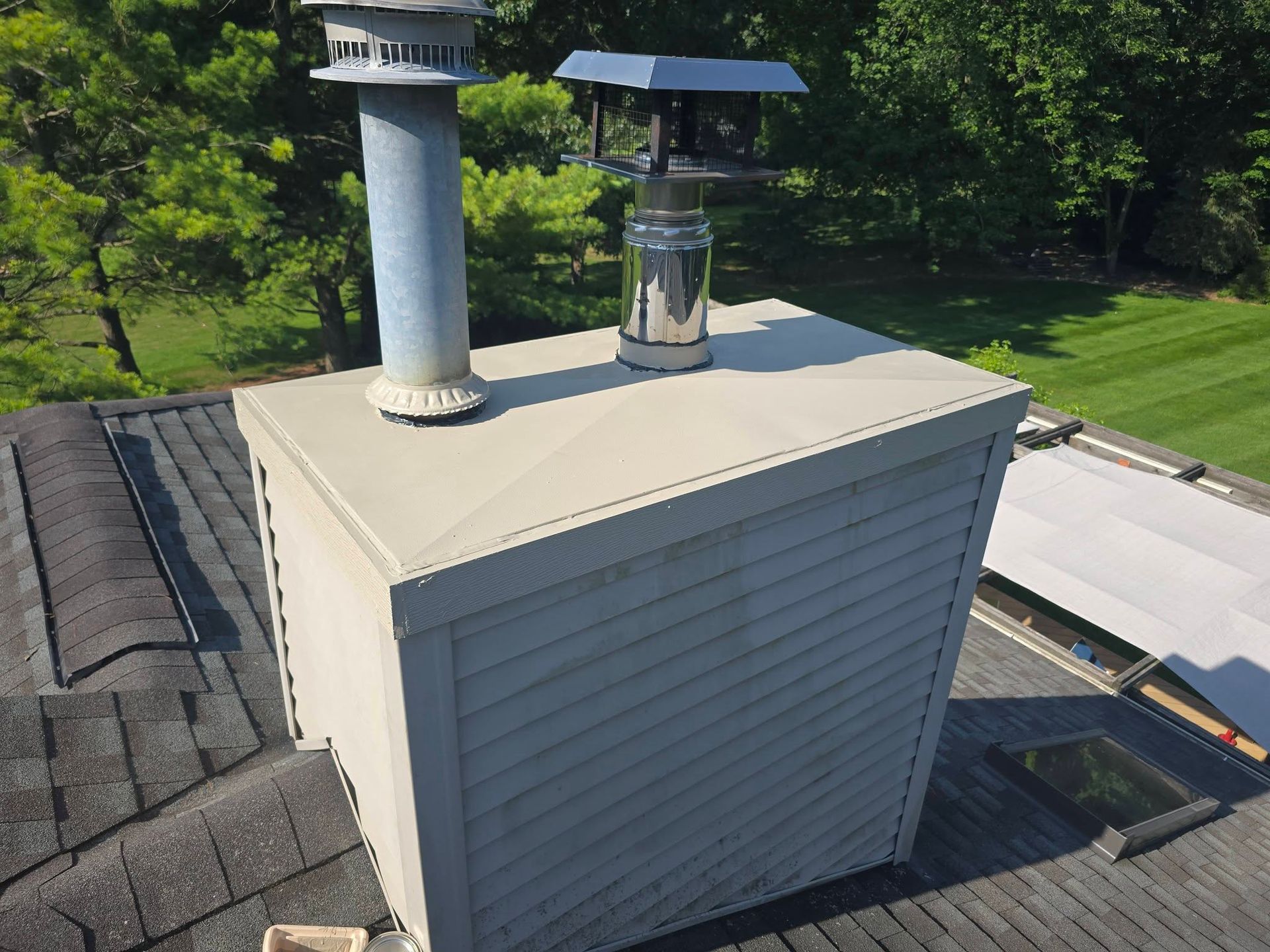 Rooftop chimney vent and metal flue on a light gray roof with trees in the background