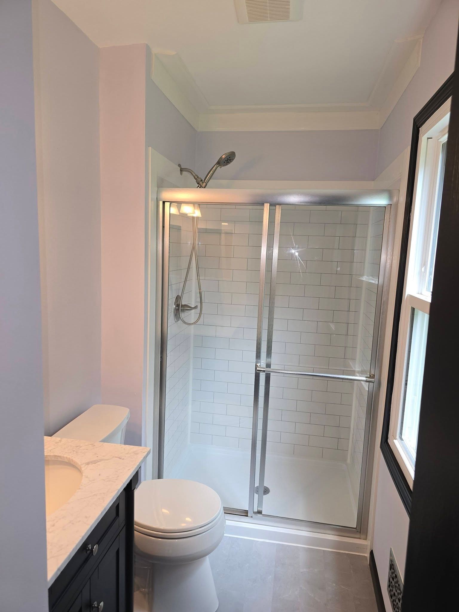 Small bathroom with glass shower, toilet, vanity, and window on the right.