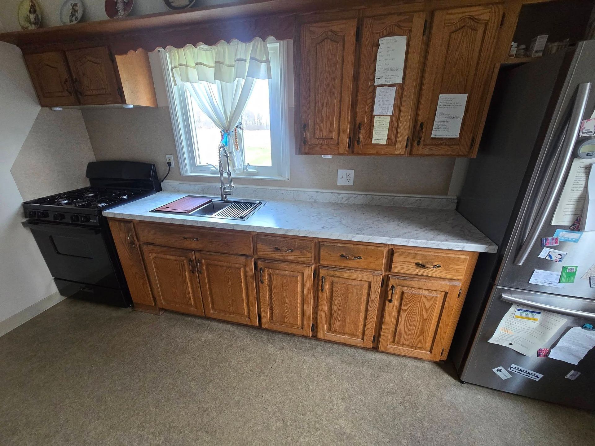 Small kitchen with wooden cabinets, white countertop, sink under window, stove left, and refrigerator right.