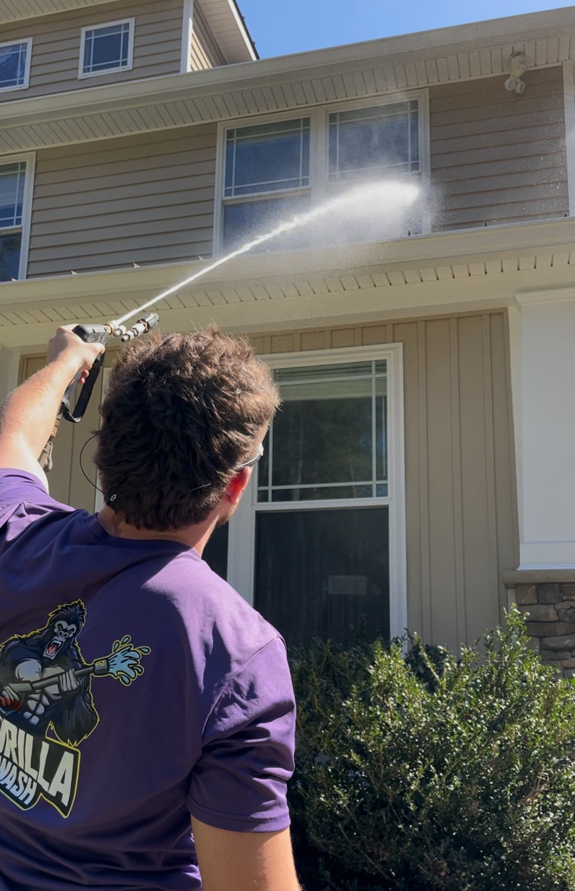 Person power-washes the exterior of a beige two-story house from the backyard.