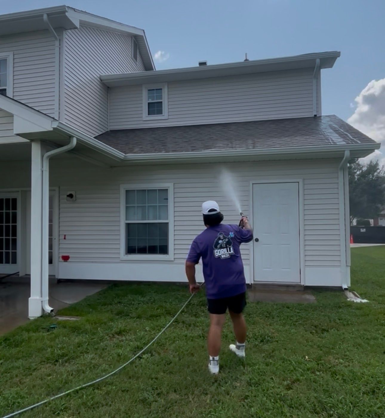 Person in a purple shirt spray-painting the side of a white house on a grassy lawn
