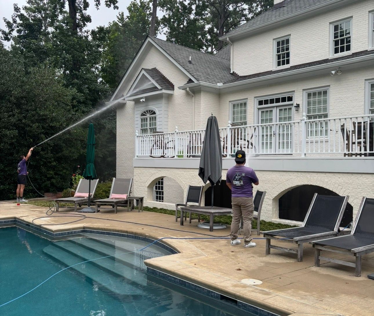 Two people pressure-wash a white house beside a backyard pool and lounge chairs.