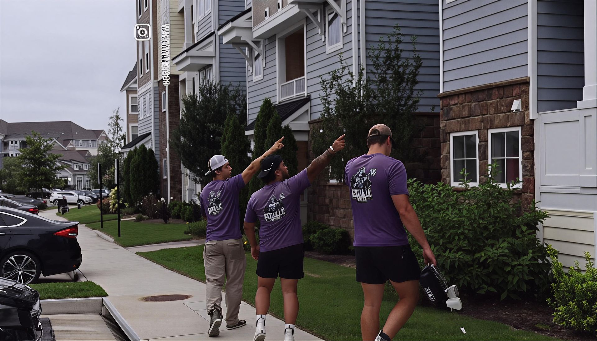 Three people in purple shirts spray watering a row of shrubs outside apartment buildings.