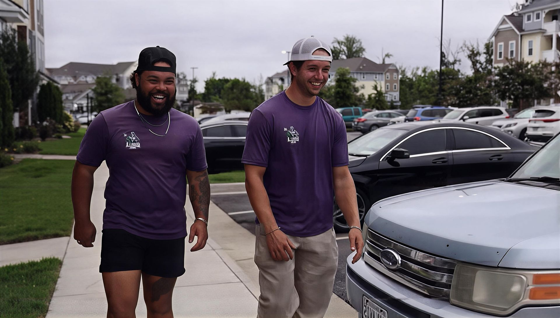 Two men in purple shirts walking on a sidewalk beside parked cars on a cloudy day