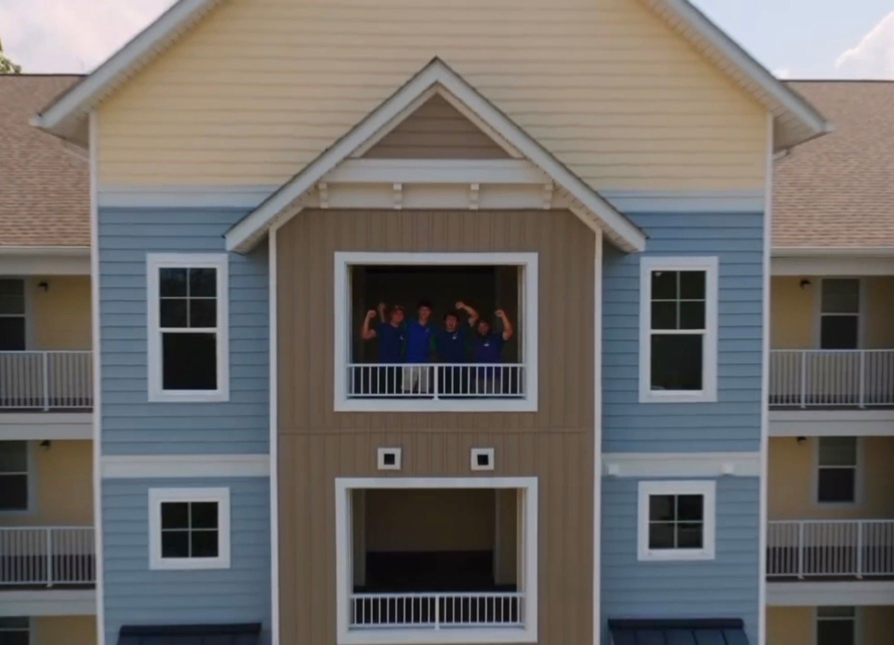 Apartment building with blue siding and balconies, with several people standing on the upper balcony
