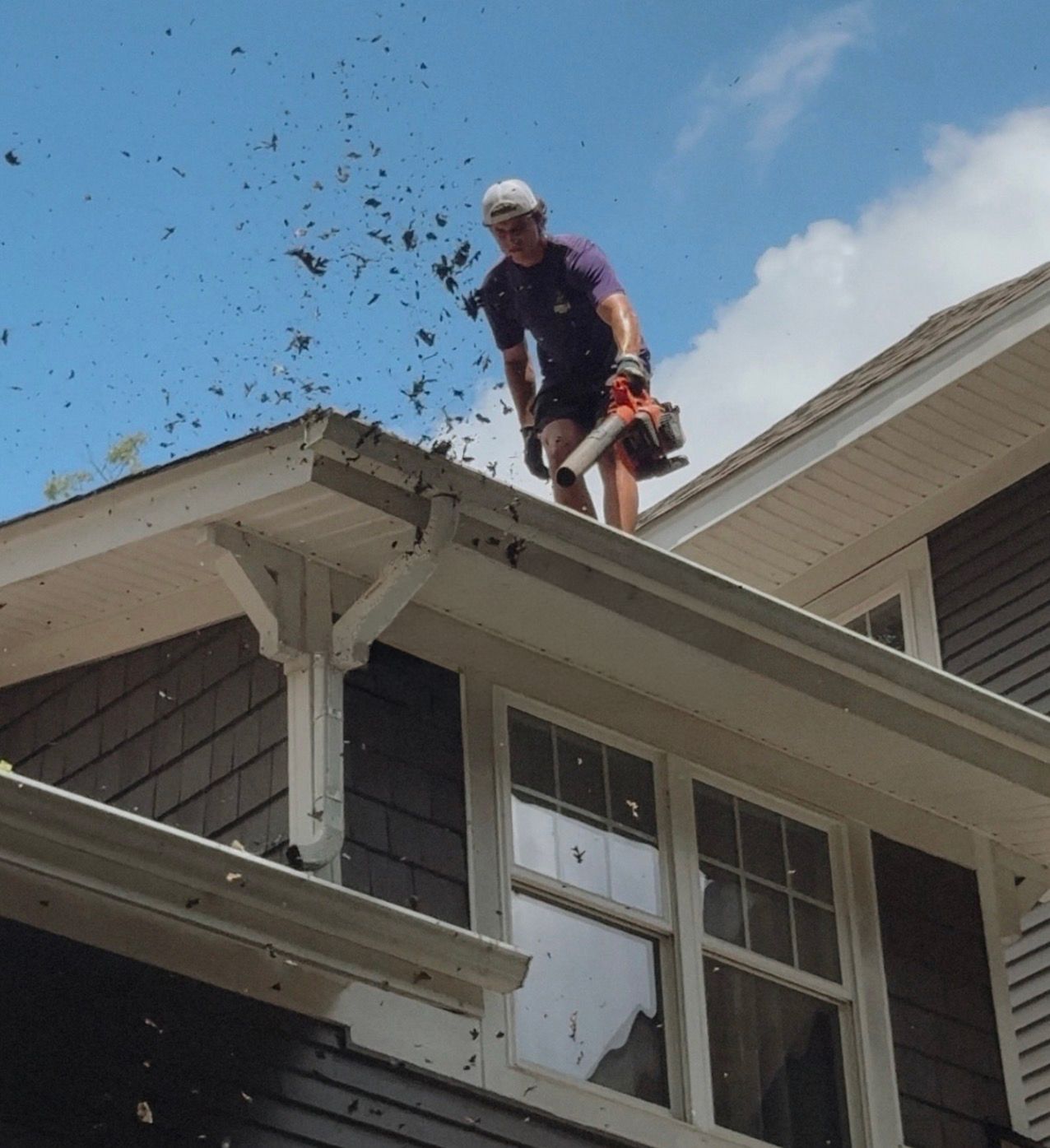 Worker using a leaf blower on a house roof, with leaves flying into the sky.