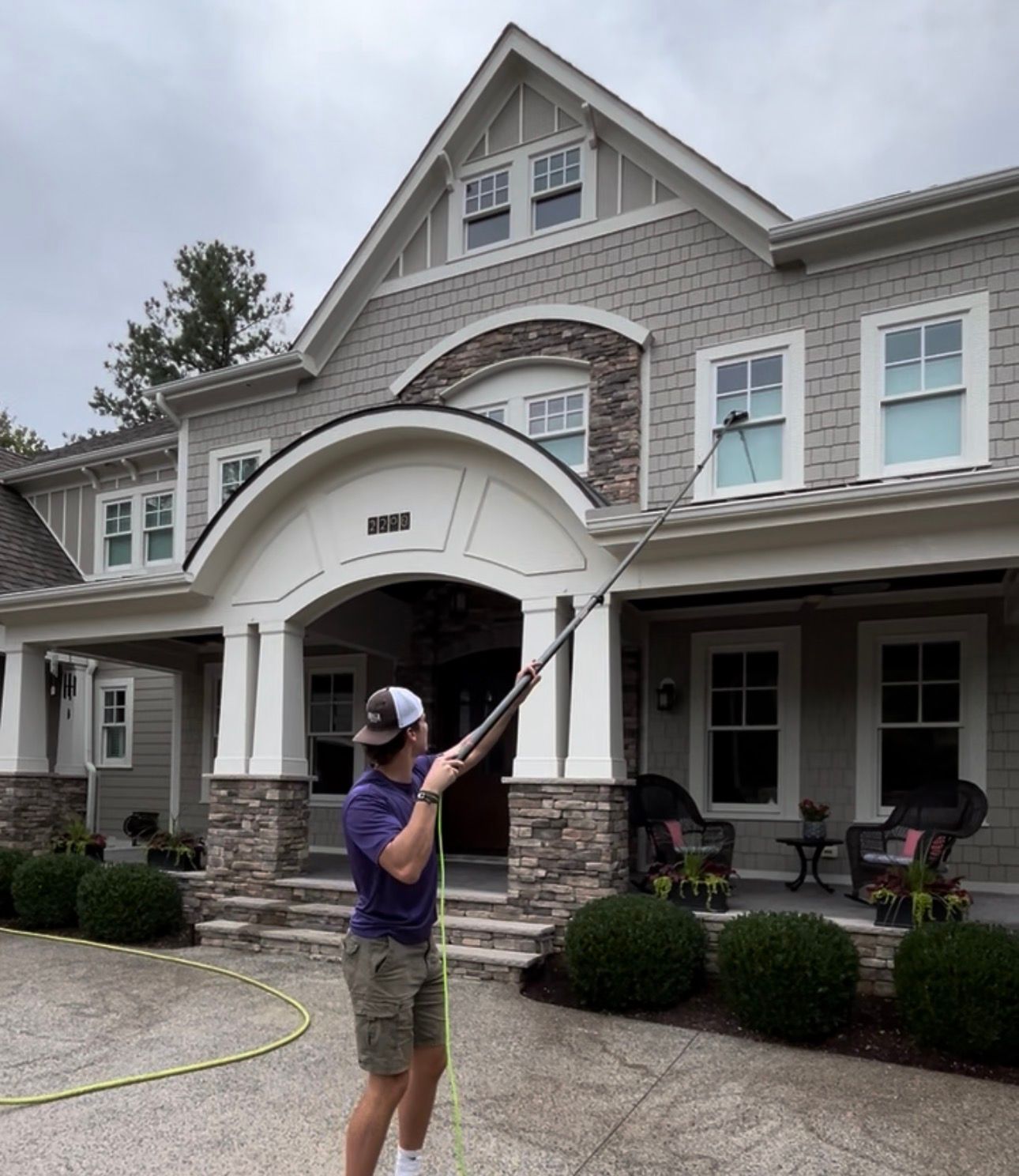 Person pressure-washing the front of a large gray house with a long wand.