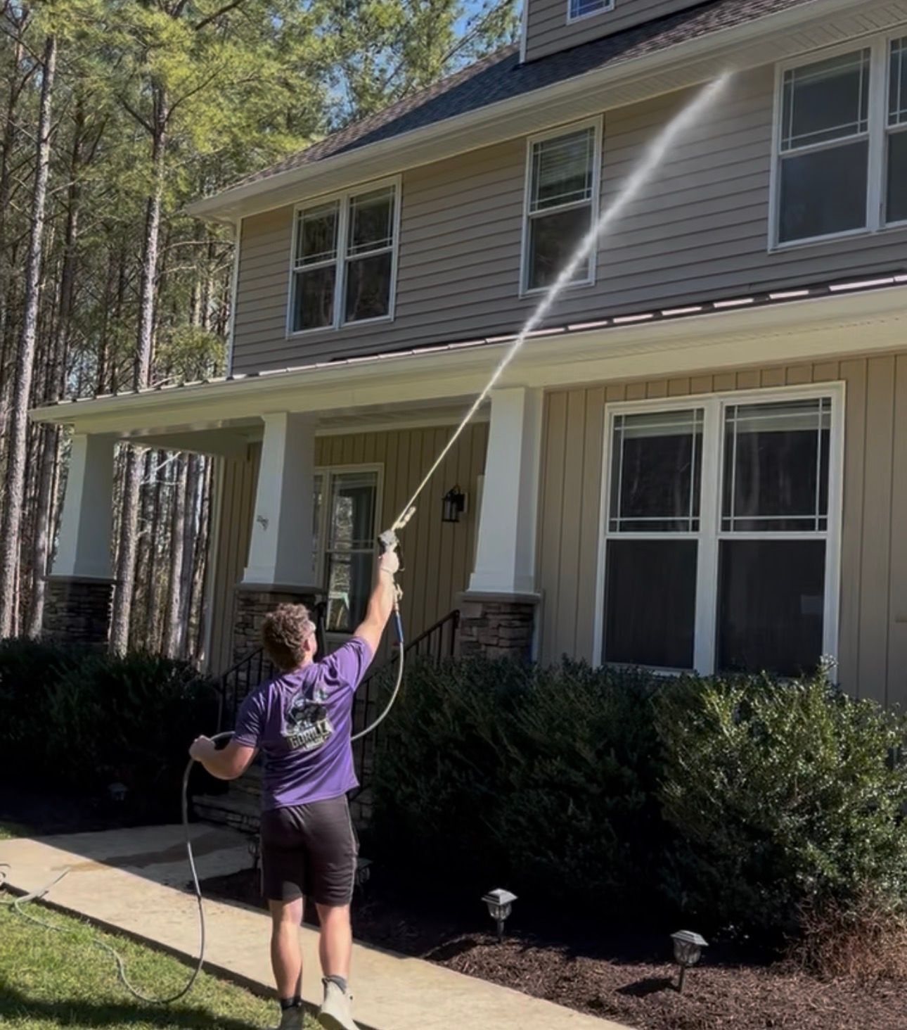 Person spraying the side of a house with a pressure washer in a suburban yard
