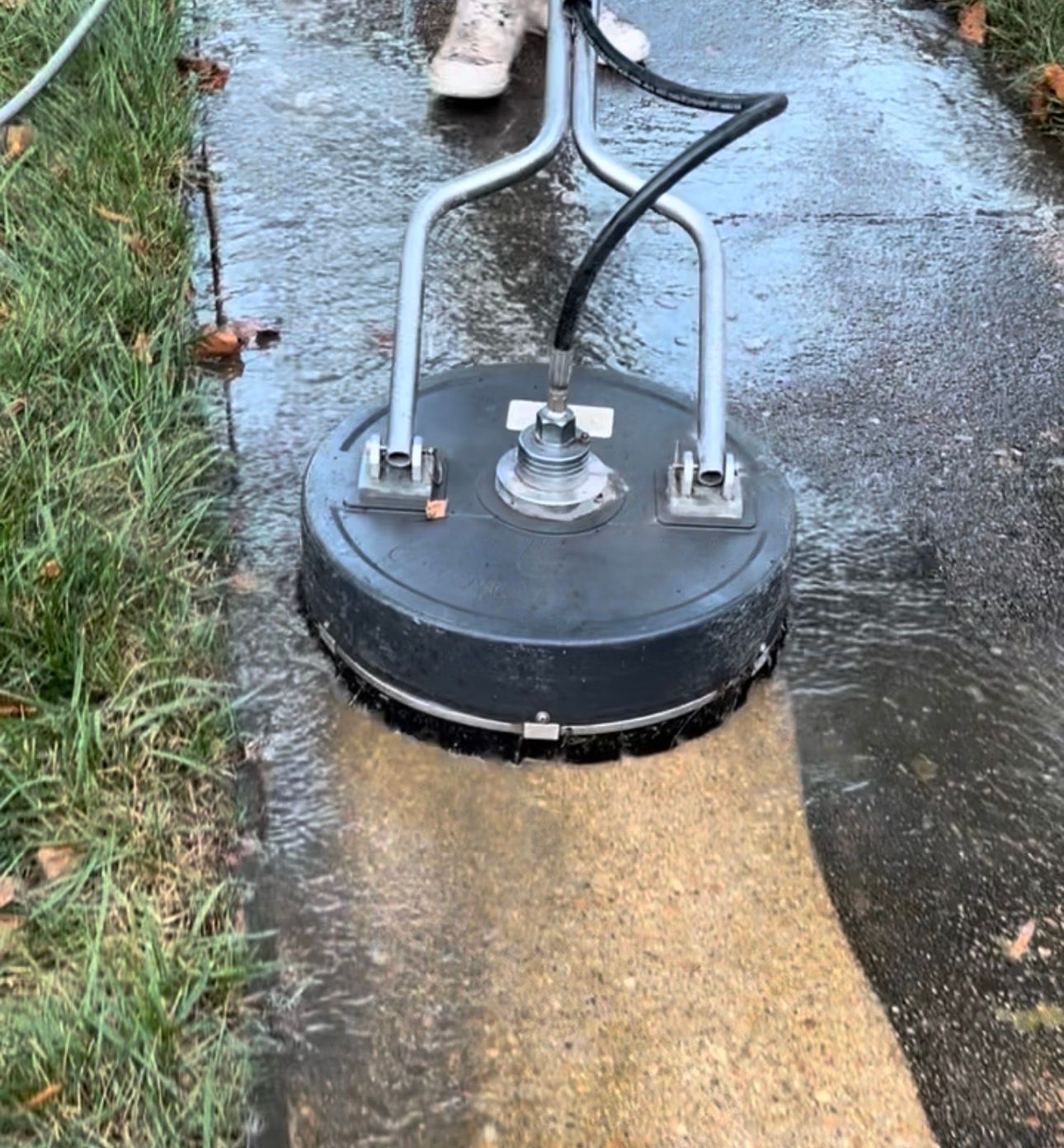 Round black cleaning machine on wet sidewalk, with hoses attached and water spraying onto the pavement.