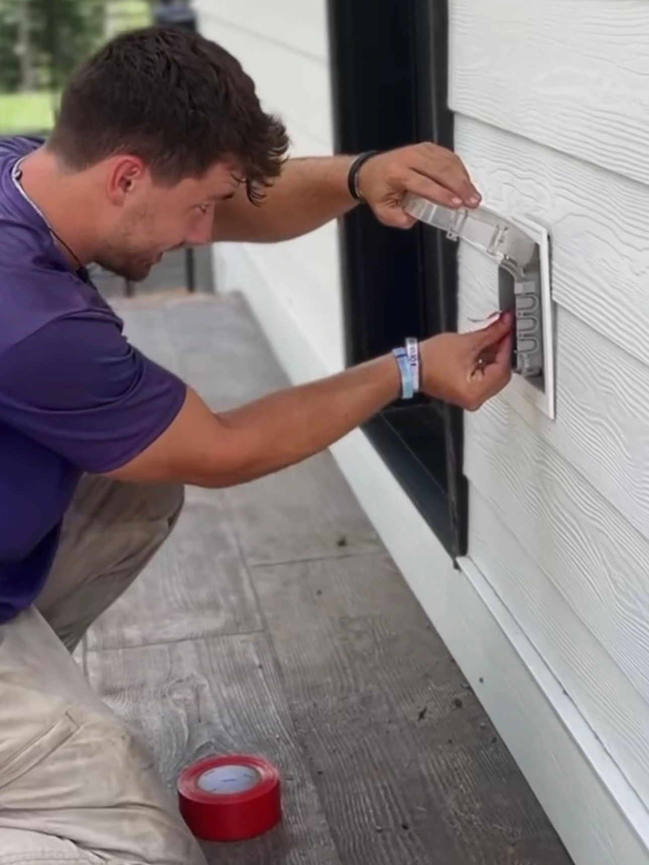Man installing a wall-mounted device on a house exterior, using a tool near a window, with red tape on the ground