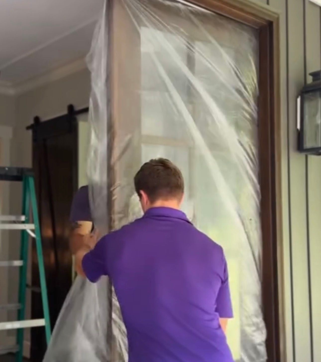 Two people wrapping a doorway in clear plastic sheeting during a home renovation.