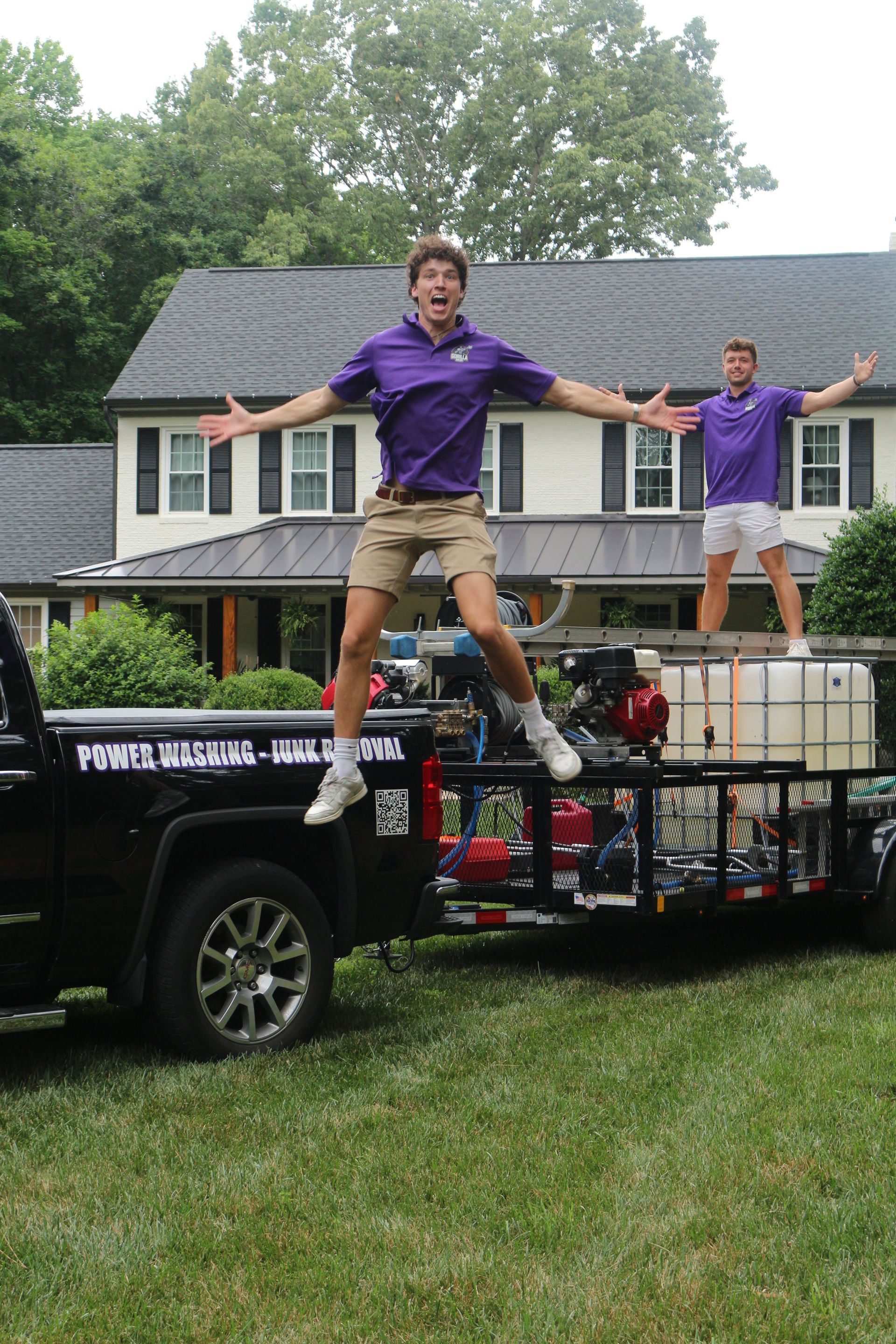 Two people balancing on a flatbed truck in a grassy yard in front of a house