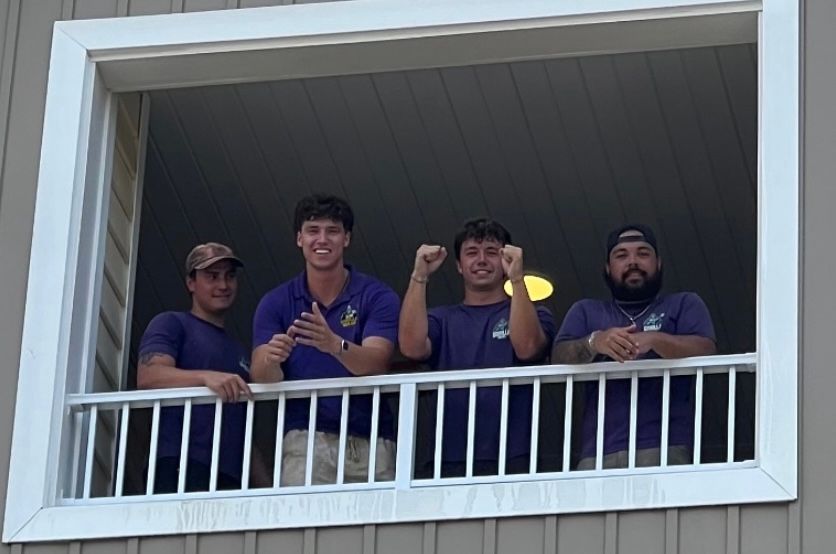 Four people leaning on a balcony railing, smiling and giving thumbs up at a window opening.