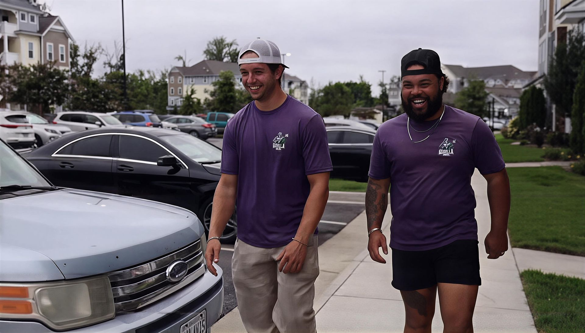 Two men in purple shirts walking beside parked cars in a suburban neighborhood