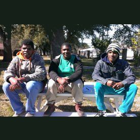 Three people sitting on a park bench. One wears a camo jacket, another a green shirt and black jacket, the third a denim jacket.