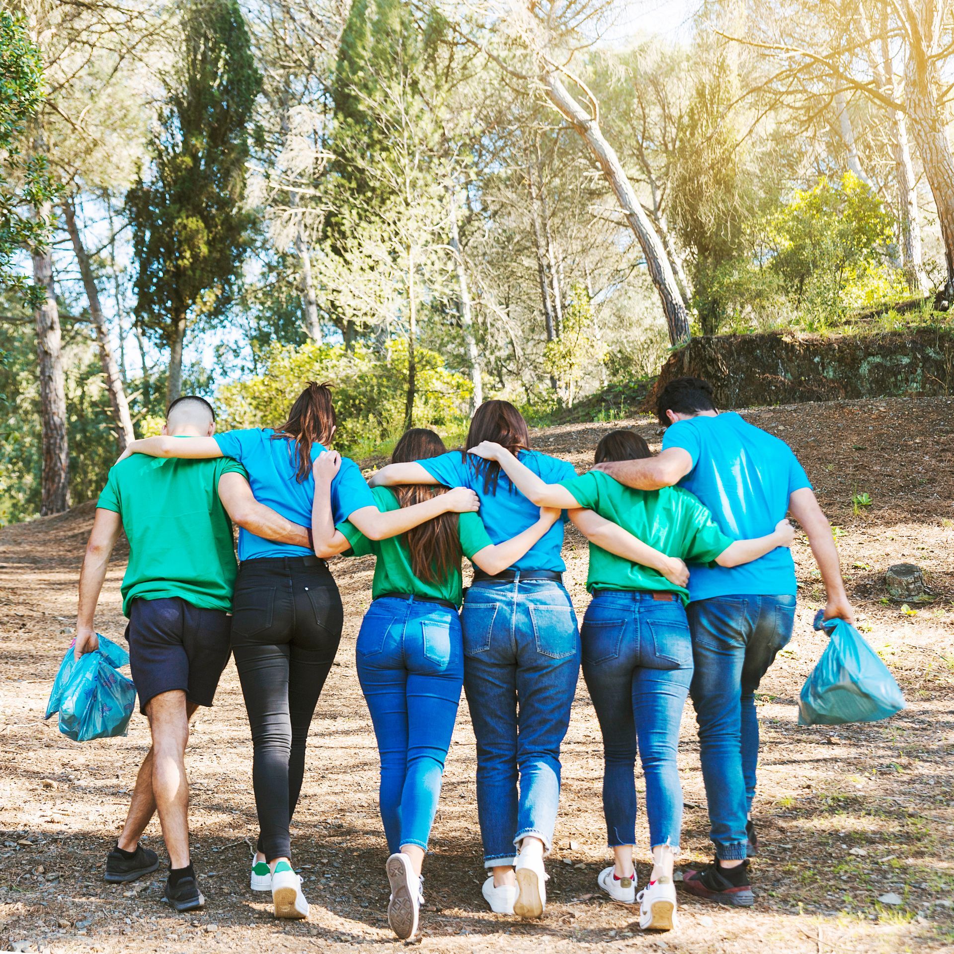 Six people with arms around each other walk through a wooded area, carrying trash bags.