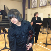 Person with cello; orchestra rehearsal, indoor gym. Conductor in background, music stands, wooden floor.