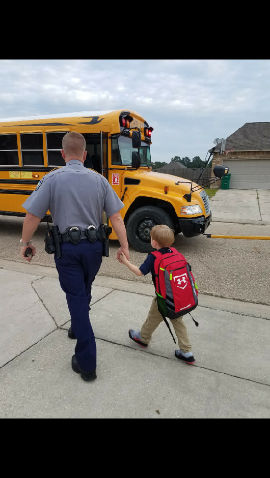 Officer holds hands with a young child, walking toward a yellow school bus on a sidewalk.